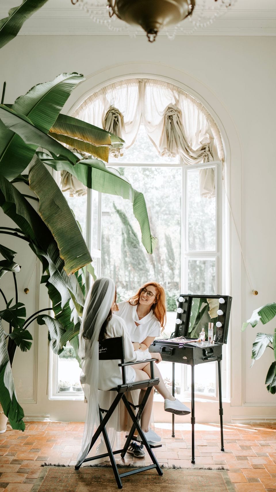 Artist seated at a lit vanity by a window, surrounded by plants